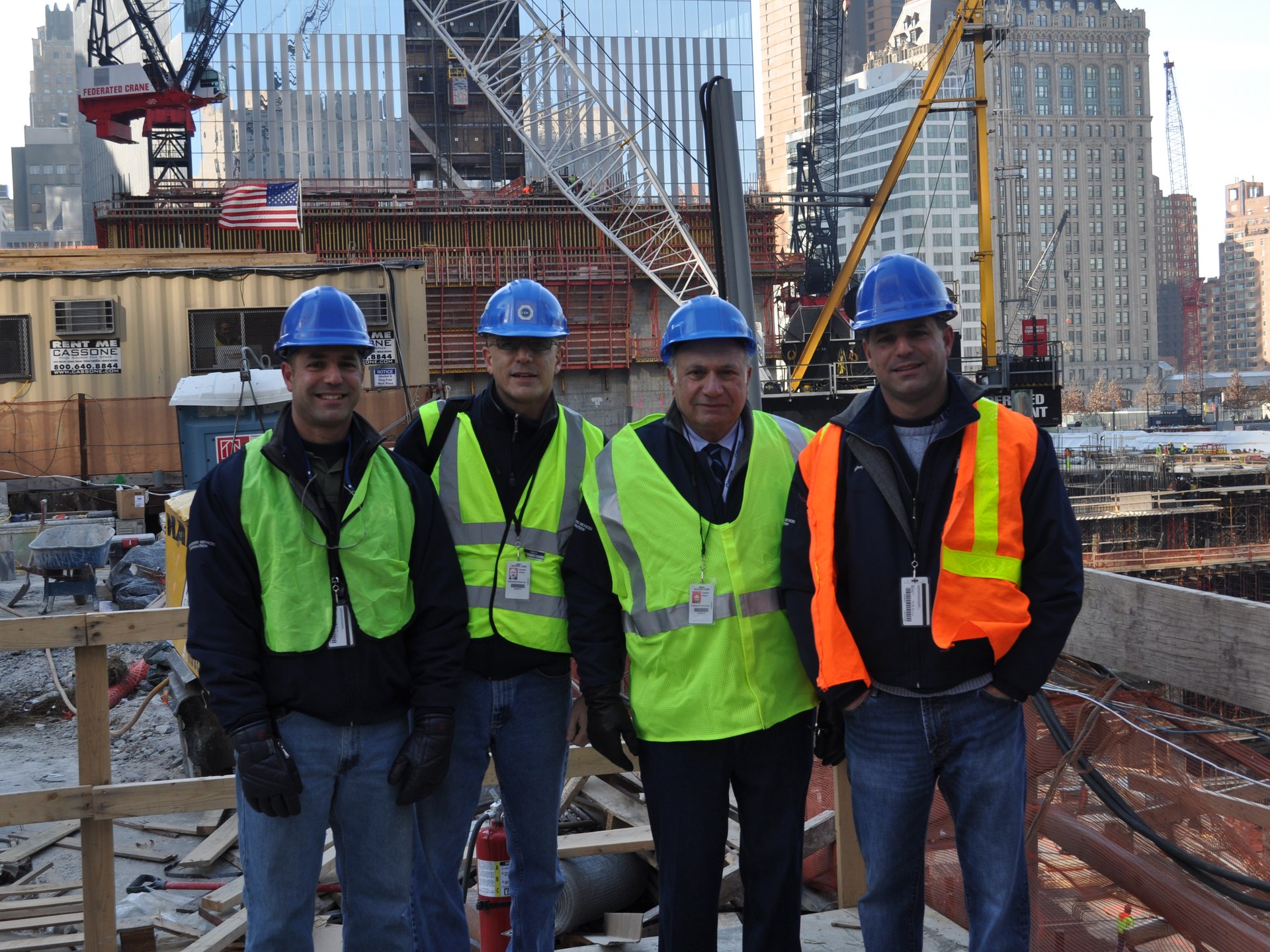 Four workers in hard hats and hi-vis vests at a concrete construction jobsite, cranes and city buildings behind