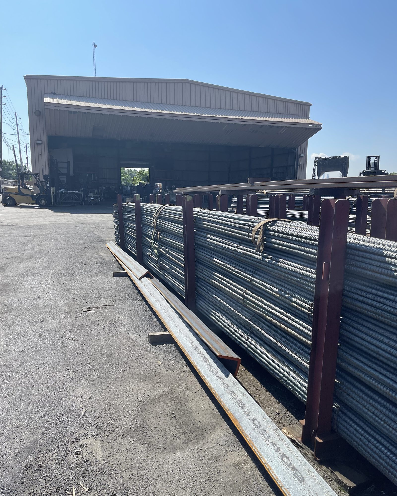 Bundles of ribbed steel reinforcing bar stacked in red steel racks in a paved yard, with an open-fronted metal warehouse building and a forklift behind them under a clear sky